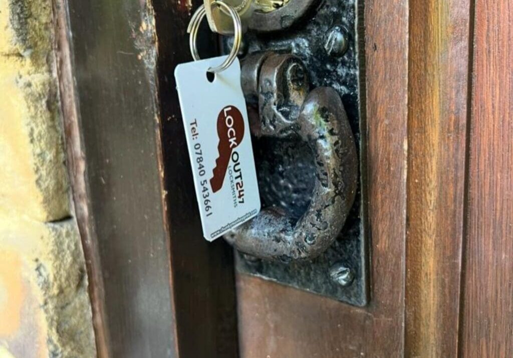 Close-up photograph showing a rim cylinder lock installed on a wooden door with a key inserted and a Lockout 24/7 locksmith tag attached