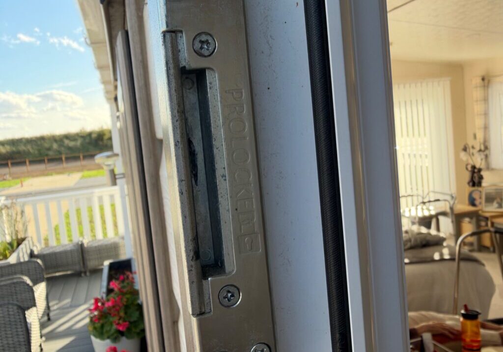 Close-up of a metal keep plate with screws fixed to a uPVC door frame at a mobile home site in Burnham-on-Crouch, with outdoor furniture and flowers visible in the background.