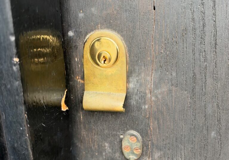 Close-up of a brass rim cylinder fitted to a black wooden front door with a handle below it in Coggeshall