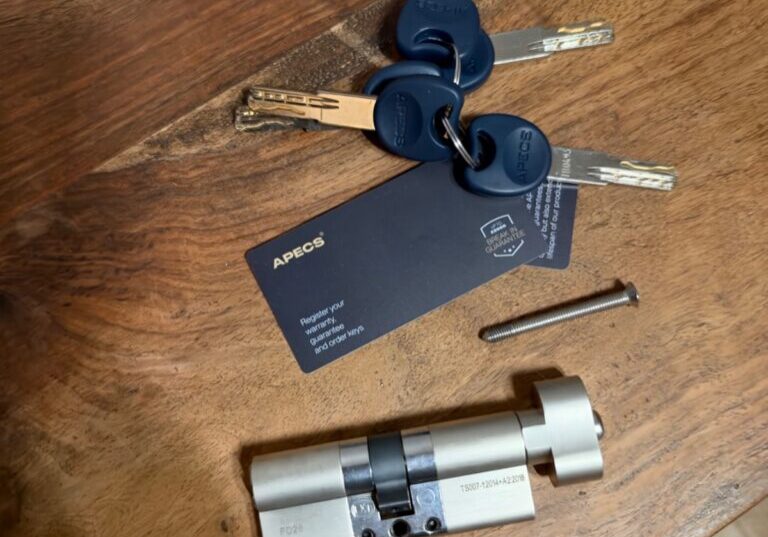 Close-up of a new silver lock cylinder with an internal thumb turn, five blue key heads and a screw, resting on a wooden surface.