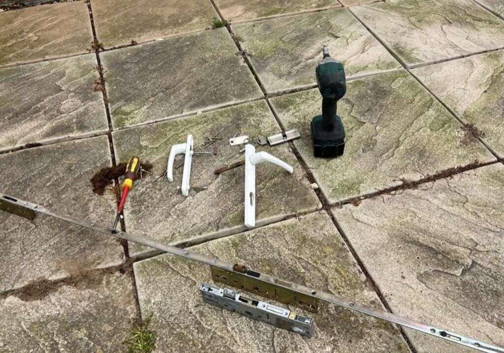 Multipoint locking gearbox, door handles, screws, and drill placed on patio slabs outside a property in Pebmarsh.